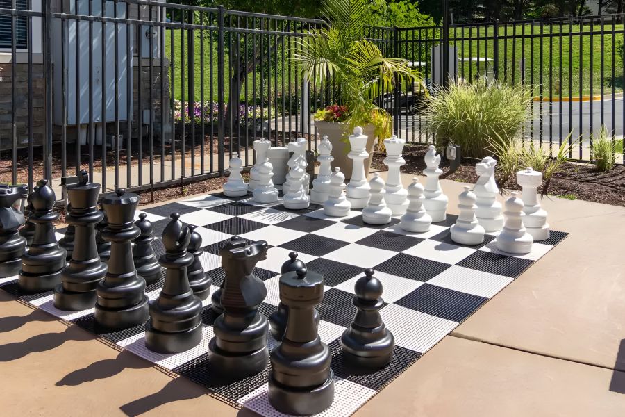 Giant outdoor chessboard with large black and white chess pieces on a patio near a fenced garden.