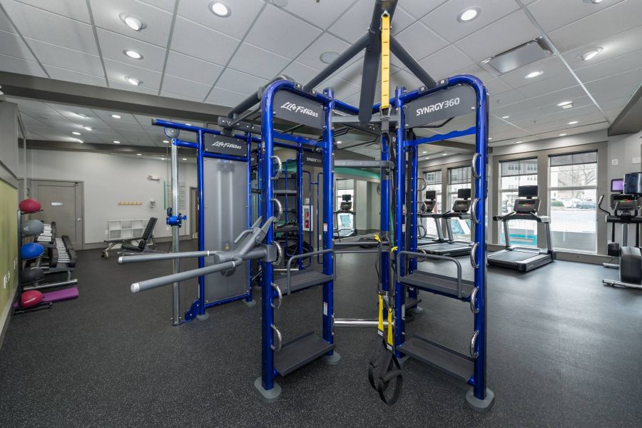 Modern gym with exercise machines, weight racks, treadmills, and large windows letting in natural light.