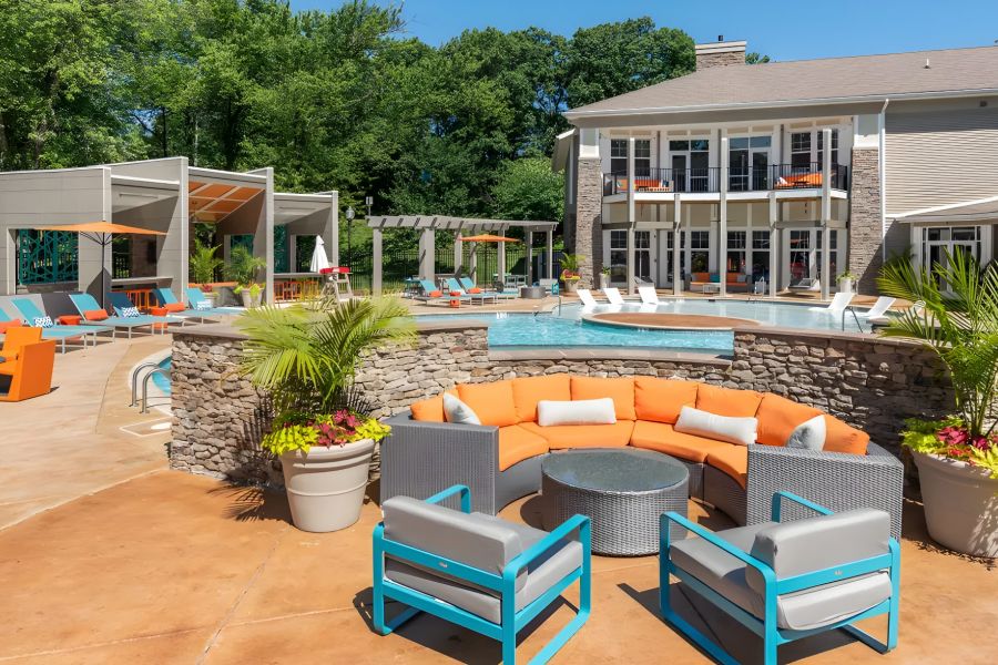 Modern outdoor pool area with orange and blue lounge chairs, patio seating, and greenery near apartments.