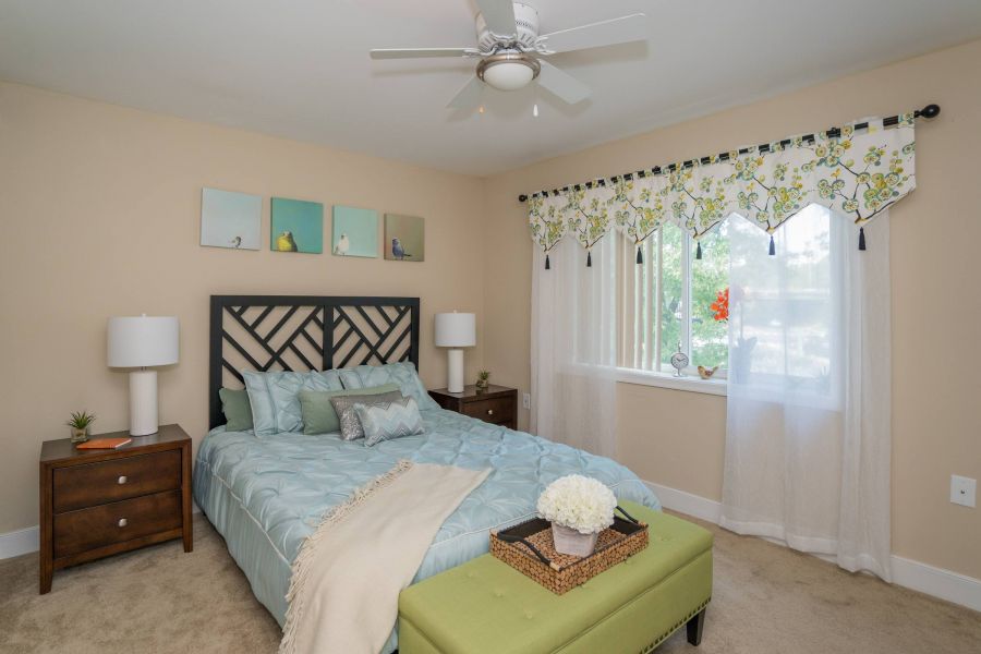 Bedroom with a bed, green bench, lamps, wall art, and a window with sheer curtains and floral valance.