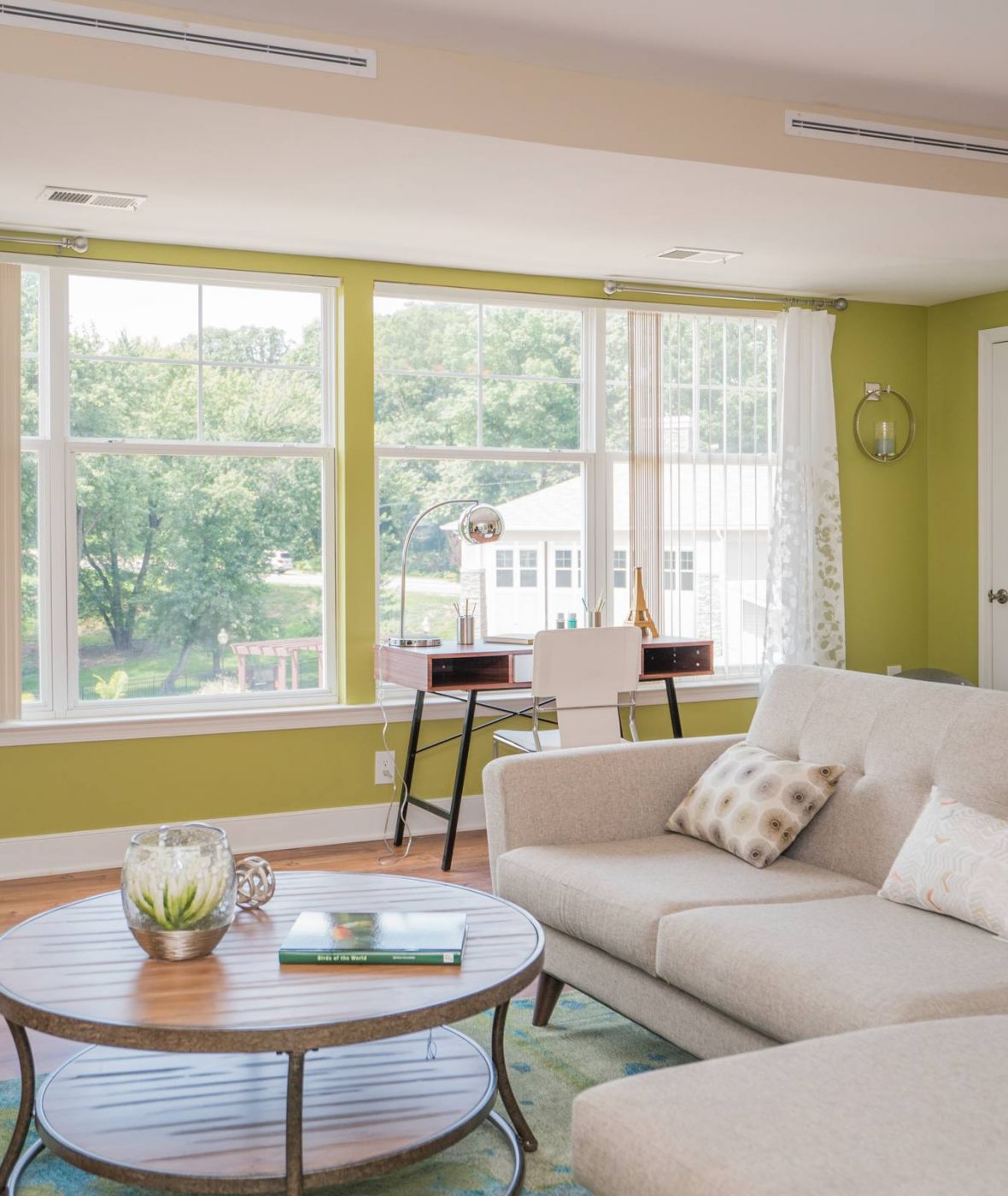 Bright living room with a beige sofa, round coffee table, and large windows letting in natural light.