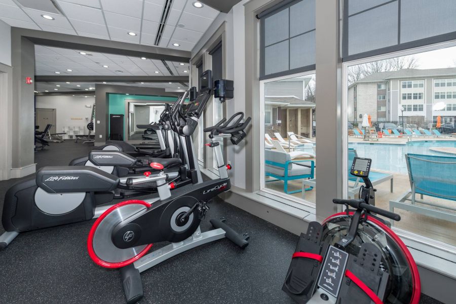 Modern gym with exercise bikes and treadmills by large windows overlooking a pool and patio area.