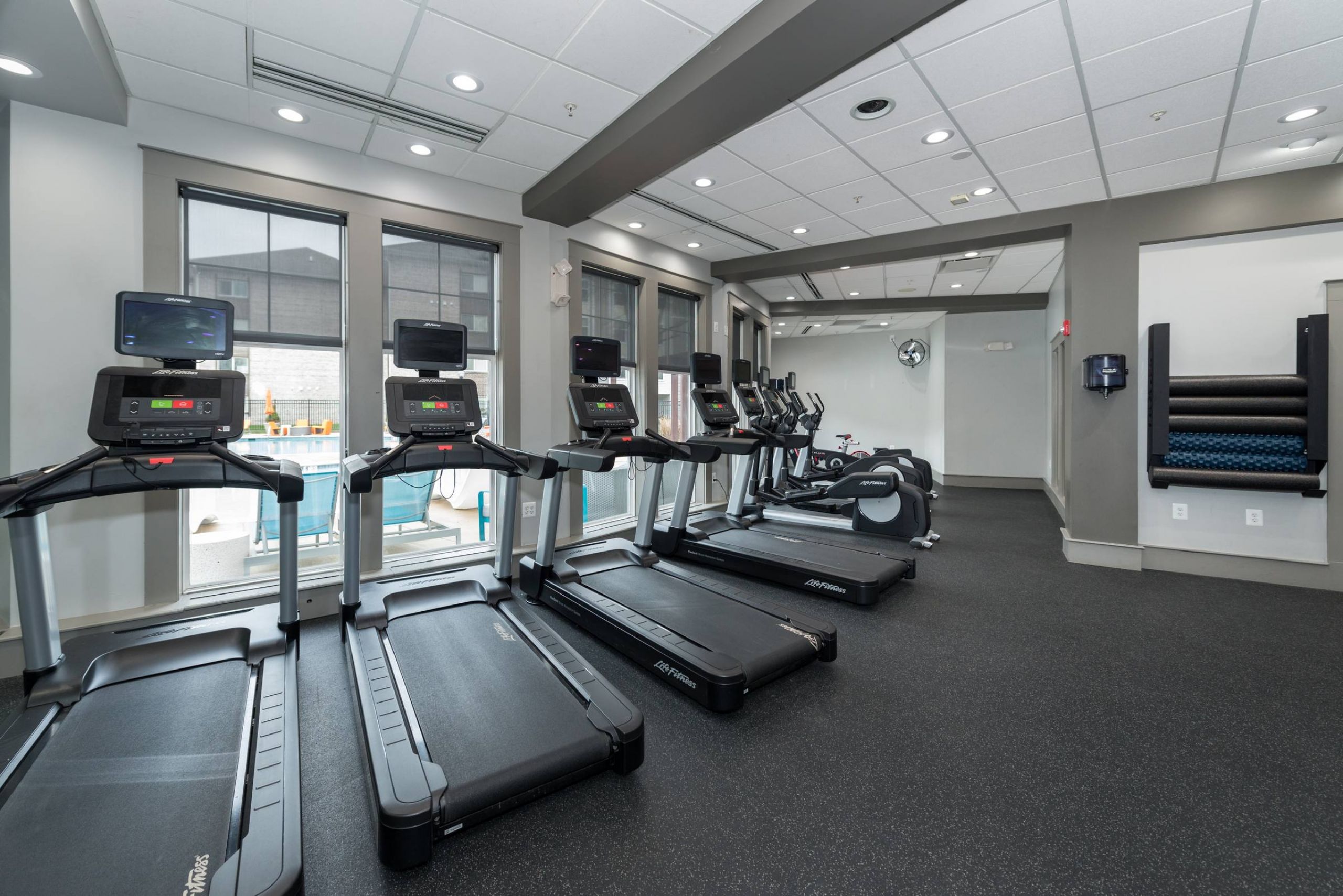 A modern gym with four treadmills, exercise bikes, large windows, and rolled exercise mats on the wall.