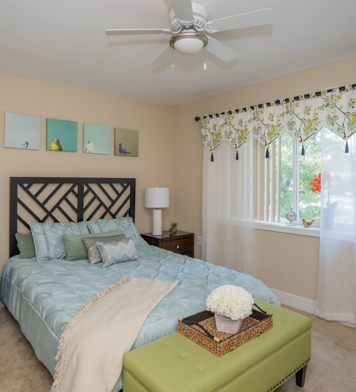 Bedroom with a bed, green bench, lamps, wall art, and a window with sheer curtains and floral valance.