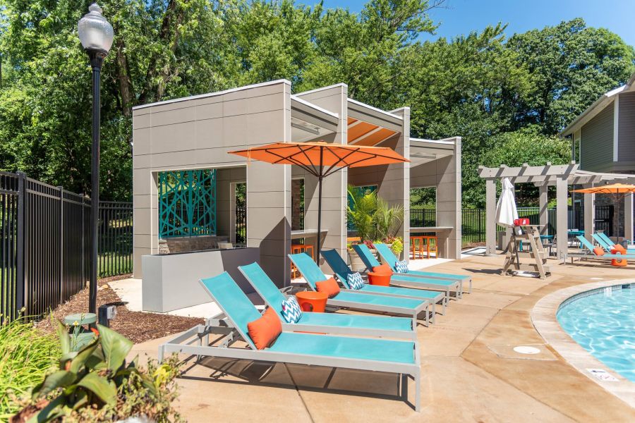 Poolside lounge chairs with orange umbrellas and cabanas on a sunny day, surrounded by greenery.